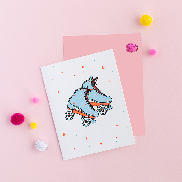 White greeting card with blue and coral/orange skates with dots and stars. There is a yellow envelope and colorful poms in the background.