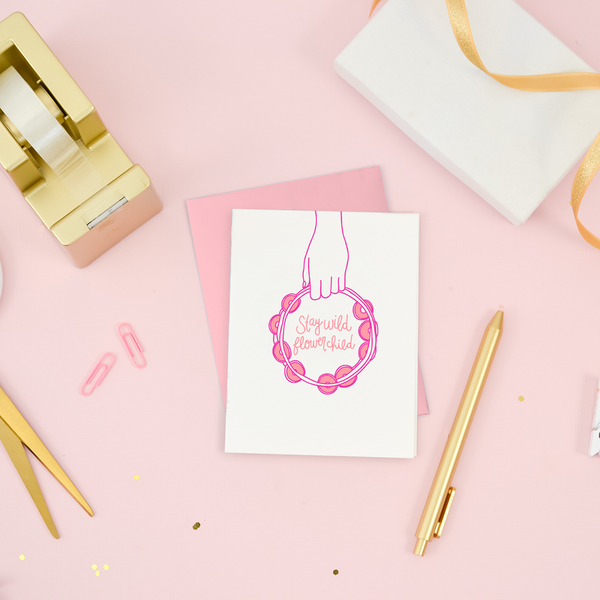Desk scene with gold desk items and a white greeting card with a hand holding a pink tambourine and the text "Stay Wild Flower Child". 