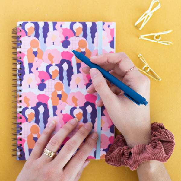 girl with pen in hand, scrunchie on wrist writing in a colorful notebook