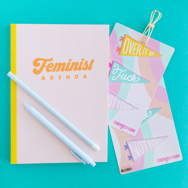 A peach "Feminist Agenda" notebook displayed with two "Over it AF" sticker sheets and a Powder blue pencil and Jotter Pen.