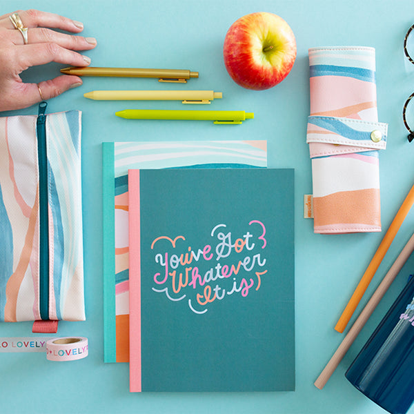 A "You've got whatever it is" notebook in a teal color, along with a notebook below it with a multicolor wave-like design. Above then notebook are three different colored Jotter pens along with a multicolored pixie pouch to the side and a Tootsie roll up pouch with the same design as the bottom notebook. All is displayed on a light blue surface.