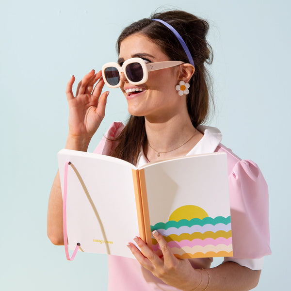 Lady in sunglasses holding up a rainbow scallop sunrise notebook.