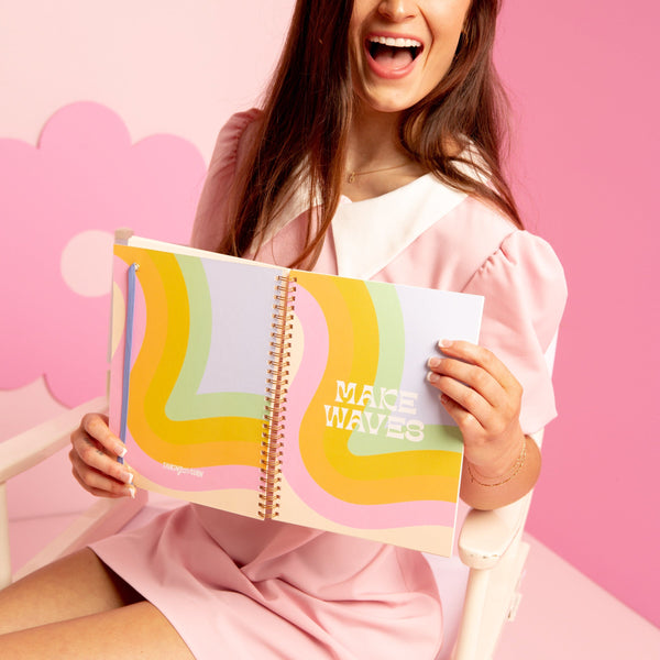 Girl in pink dress holding up a rainbow waves colorful notebook.