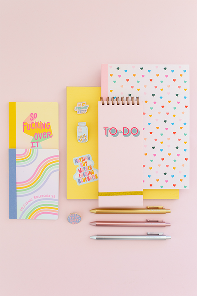 stack of different sized notebooks with different patterns, metallic pens and enamel pins