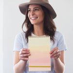 A happily smiling young woman holding a taskpad with a soft rainbow gradient in front of her.