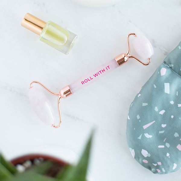 A light pink Face Stone Roller with rose gold accents. The phase "Roll with it" is printed on in pink lettering. Face Stone roller is displayed with a cosmetic product and a terrazzo speckled Eye Mask.