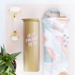 A white Face Stone Roller with gold accents displayed next to a gold colored tumbler with the phrase "Is it Friday Yet?" printed in light purple lettering. Next to tumbler is a multicolored pastel neck wrap. All items are on a light pink and white background.