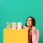 Colorful mini notebook set standing up in a line with a girl smiling next to them. The note books are rainbow scalloped, column of daisies, and "what a time to be alive" text.  