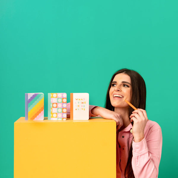 Colorful mini notebook set standing up in a line with a girl smiling next to them. The note books are rainbow scalloped, column of daisies, and "what a time to be alive" text.  