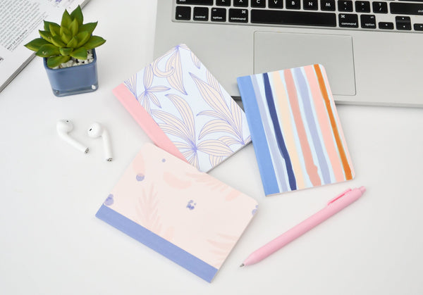 three mininotebooks next to a laptop, airpods, a plant, and a pink jotter pen. One notebook has periwinkle leaves, one has stripes, and one is a watercolor