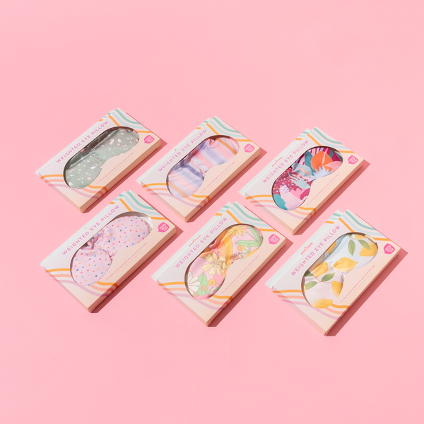 a weighted eye mask with yellow lemons in toots colorful packaging with a group of all eye pillow patterns on a pink background.