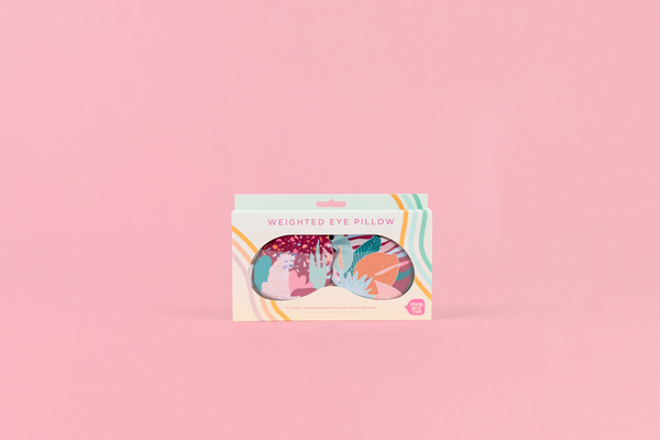 Weighted eye mask with collage imprint with burgundy, peach, light blue, orange and teal in toots colorful packaging on a pink background.