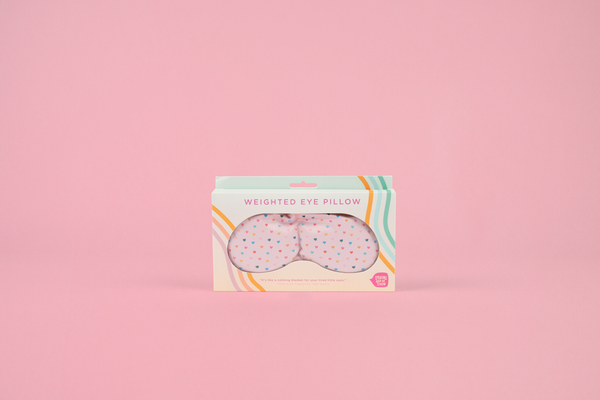 a pink weighted eye mask with assorted color tiny hearts in toots colorful packaging on a pink background.