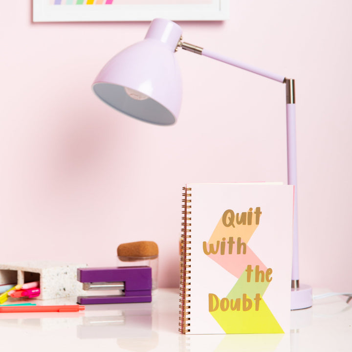 light pink binder in golden text saying 'Quit with the Doubt' each word being down lit in brown, pink, dark green and light green with a golden elastic band to close the planner. next to a lavender lamp, purple stapler and a large pen cup with multiple colored pens.