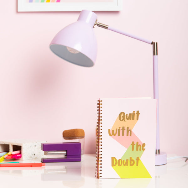 light pink binder in golden text saying 'Quit with the Doubt' each word being down lit in brown, pink, dark green and light green with a golden elastic band to close the planner. next to a lavender lamp, purple stapler and a large pen cup with multiple colored pens.