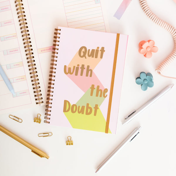 light pink binder in golden text saying 'Quit with the Doubt' each word being down lit in brown, pink, dark green and light green with a golden elastic band to close the planner. surrounded by jotter pens, paper clips and flower clips.