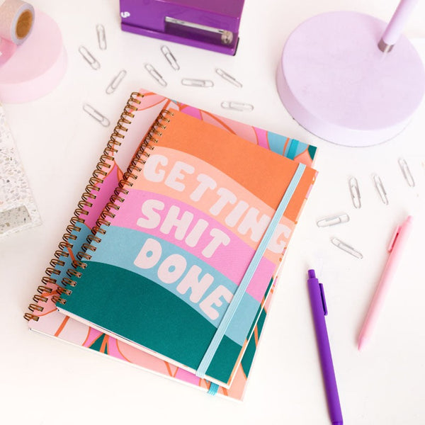 small goal getter planner with five waves in green, blue, pink, peach and orange, the words 'getting shit done' in white fluffy text at the front. surrounded by paper clips, two pen jotters, purple stapler and a lavender lamp.