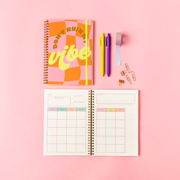"Don't Ruin My Vibe" Perpetual Planner - Goal Getter Lite - Talking Out Of Turn. Orange and pink checkered pattern. Open and closed undated planner. opened on monthly calendar with jotter pens, wash-tape, and paper clips on a pink background.
