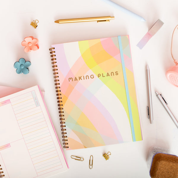 a perpetual planner with pastel multi-color waves with text saying 'making plans' and a bright blue elastic ban to keep the planner closed. surrounded by pens, paper clips and flower hair clips.