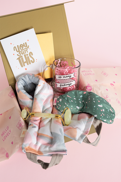 a collection of self care items packaged in a gold box including a neck wrap and an eye mask, a clear glass mug with "So many feelings" written on it. A card that says "You Got This", and a face stone roller 