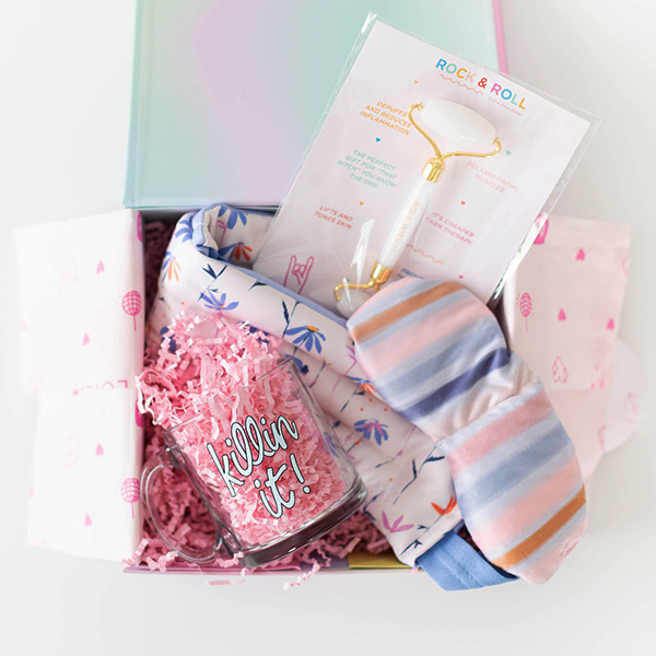 A gift set containing a clear glass mug with Killing It written in blue, a neck wrap with small flowers, a striped eye mask, and a face roller all in pink crinkle paper and packaged in a colorful box. 
