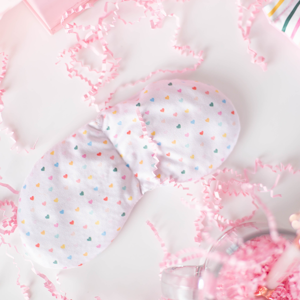 A light pink weighted eye mask with multi colored hearts printed all over. There is a clear glass mug in the bottom right corner and light pink crinkle paper scattered all over. 