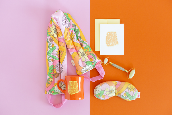 flat image of a collection of self care items including a neck wrap and an eye mask in a pink floral and swirl print, an orange diner style mug and  a card that say all the feels, and a face stone roller 