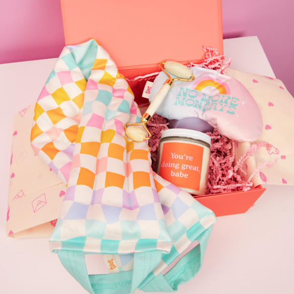 Carnival checkers weighted neck wrap and a "no more mondays" weighted eye pillow with a face roller and a "you're doing great, babe" candle in a orange box on a white table and a pink background.