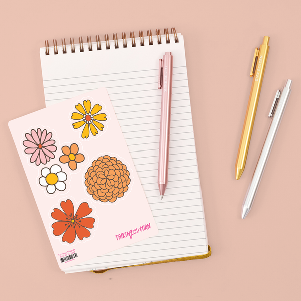 four cute stickers of various flowers on top of notebook with three metallic pens