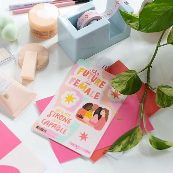 A "The Future is Female" sticker set with star stickers, a "You are strong and capable" sticker, and a sticker with four women hugging each other. Colors of sticker are yellows, oranges, and pinks. Sticker Set is displayed with a powder blue desk set piece, a plant, and some cosmetic products on a white surface.