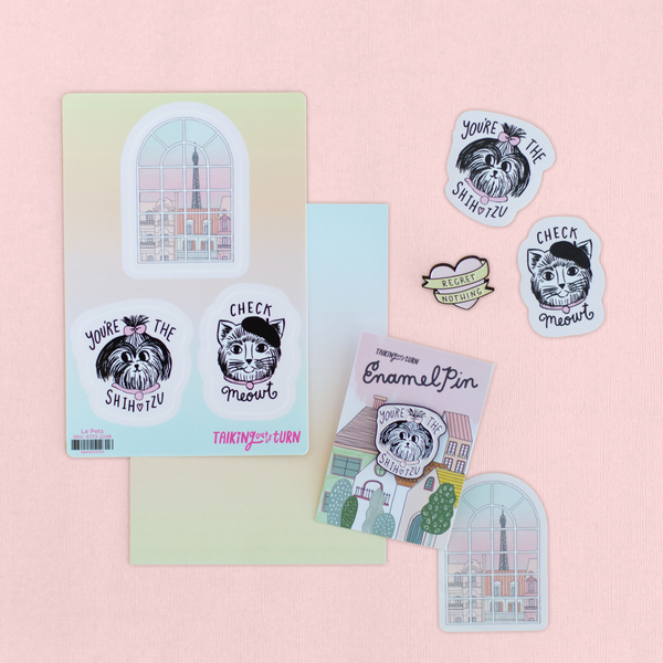 A "Le Pets" sticker set with a dog sticker and a cat sticker, along with a window looking out of Paris sticker. A "You're the Shihtzu" enamel pin is also displayed, all in front of a light pink background.