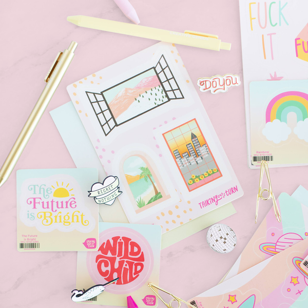 A "The Perfect View" sticker with three different city view stickers, displayed with a "Future is Bright" sticker, a "Wild Child" sticker and a "Rainbow" sticker. Enamel Pins and Jotter Pens are also displayed, all in front of a pink background.
