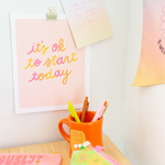 An "It's ok to start today," print with multicolored cursive lettering and an orange pastel background. Displayed on a white wall with a note and flower stickers, along with an orange "All the feels" mug sitting on a desk in front with a Flower power pixie pouch and some Jotter pens.