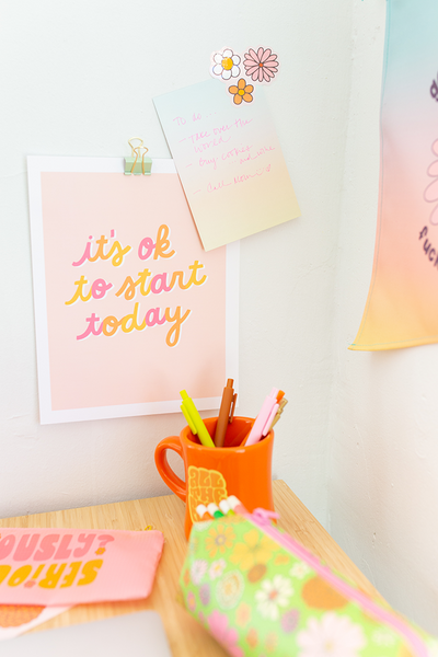 An "It's ok to start today," print with multicolored cursive lettering and an orange pastel background. Displayed on a white wall with a note and flower stickers, along with an orange "All the feels" mug sitting on a desk in front with a Flower power pixie pouch and some Jotter pens.