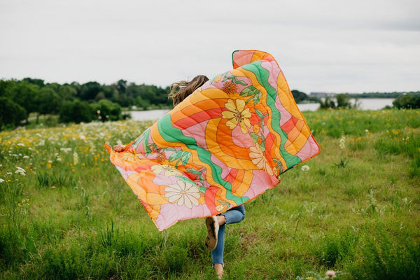girl running in field of flowers holding a blanket behind her with big flowers and rainbow swirls.