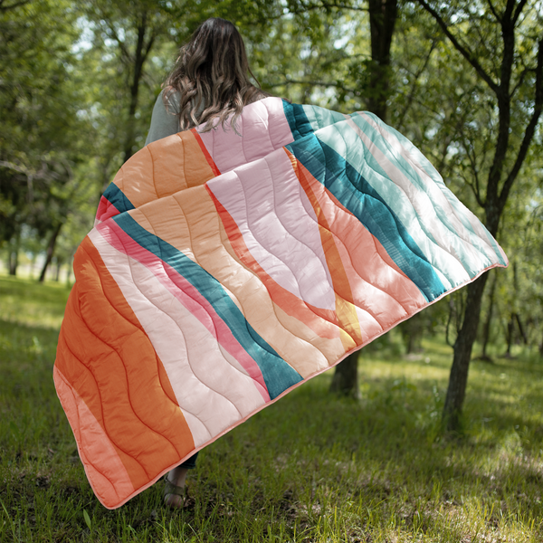 woman running in a field holding puffy quilted blanket in a pastel and teal swirl pattern with solid teal on the backside