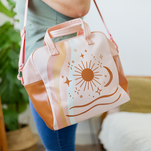 woman in olive green shirt and blue jeans holding a rainbow flow skate bag on her shoulder with sun moon stars and waves designs in pastel colors