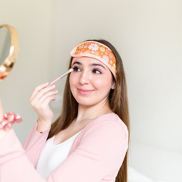 A woman wearing the Flower Power spa head wrap, holding a makeup brush near her eye and a mirror facing herself. The spa head wrap has a light pink trim, and a retro style flower pattern with an orangey-brown background. 