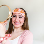 A woman wearing the Flower Power spa head wrap, holding a makeup brush near her eye and a mirror facing herself. The spa head wrap has a light pink trim, and a retro style flower pattern with an orangey-brown background.