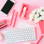 Overhead shot of an Apple keyboard, an iPhone, neon coral stapler, pens, tape dispenser, and pen cup with white flowers scattered across a pink desk.