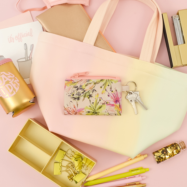 Tropical Mess Penny Key Ring is a coin purse key ring in an abstract floral pattern. Displayed with a Day Break Dumpling totebag, a gold colored desk set, and other items in front of a pink background.