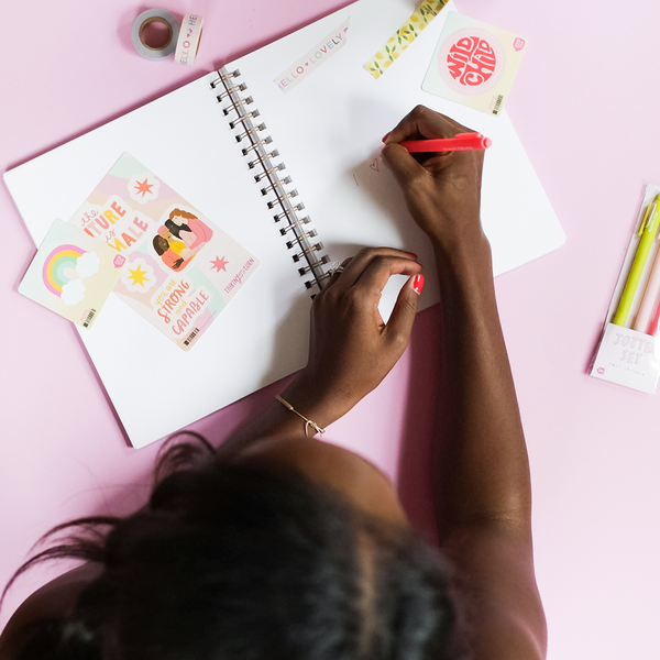 a girl writing in a notebook with a cute pink, yellow, teal, and blue rainbow sticker on top