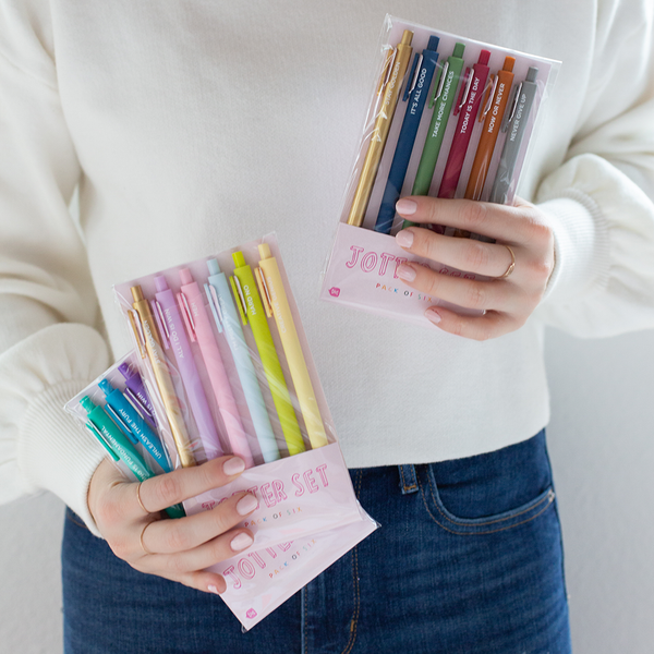 A person holding different multicolored Jotter sets. On the left, the person is holding a bright colored set and a pastel colored set. On the right, person is holding a darker set.