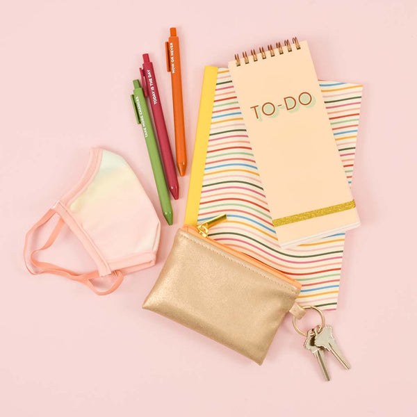 A metallic gold penny pouch with a key ring surrounded by a pastel face mask, jotter pens, a notebook and a to-do taskpad.