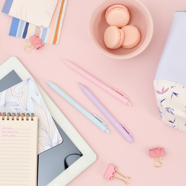 Powder blue, blush pink, and lilac Jotter Pens displayed on a pink surface and surrounded by mini notebooks, Tweedle dum pouch, a task pad, and other items.