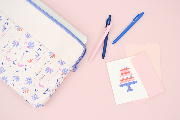A white greeting card with a layered birthday cake on a cake stand and 3 candles on top. There is a magic sprigs laptop sleeve and three jotter pens on a pink background. 