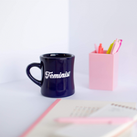 Navy blue diner funny coffee mug with Feminist written in pale pink hand lettered font next to a planner and task pad