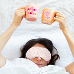 A woman laying in bed with a weighted eye mask with a bunch of colorful tiny hearts on with mugs help above her head in peach and orange with sayings on them.