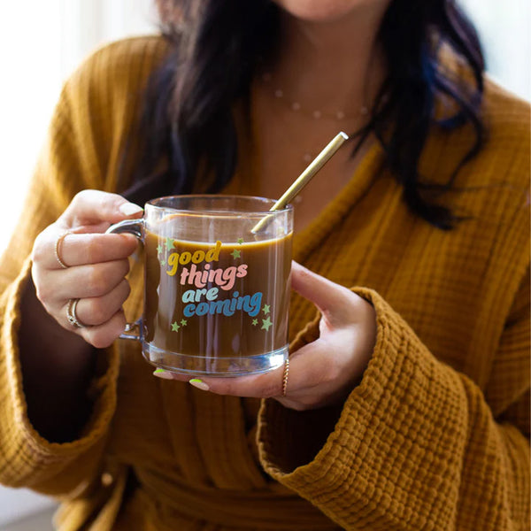 Woman in robe holding a clear glass mug with the phrase "good things are coming" printed on.