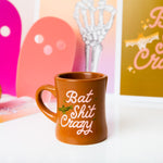 Camel color diner mug with "Bat Shit Crazy" written on the front in pink font, with illustration of bat and olive green minimalist sparkle-stars surrounding text.